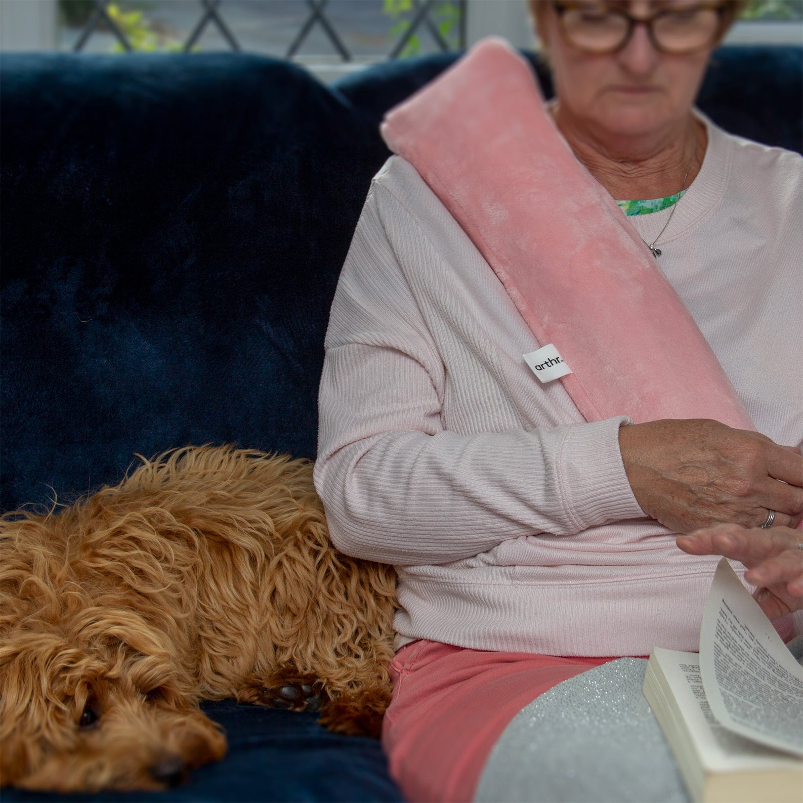 Long hot water bottle in rose quartz pink being worn by a woman sitting on a sofa with her dog and reading a book